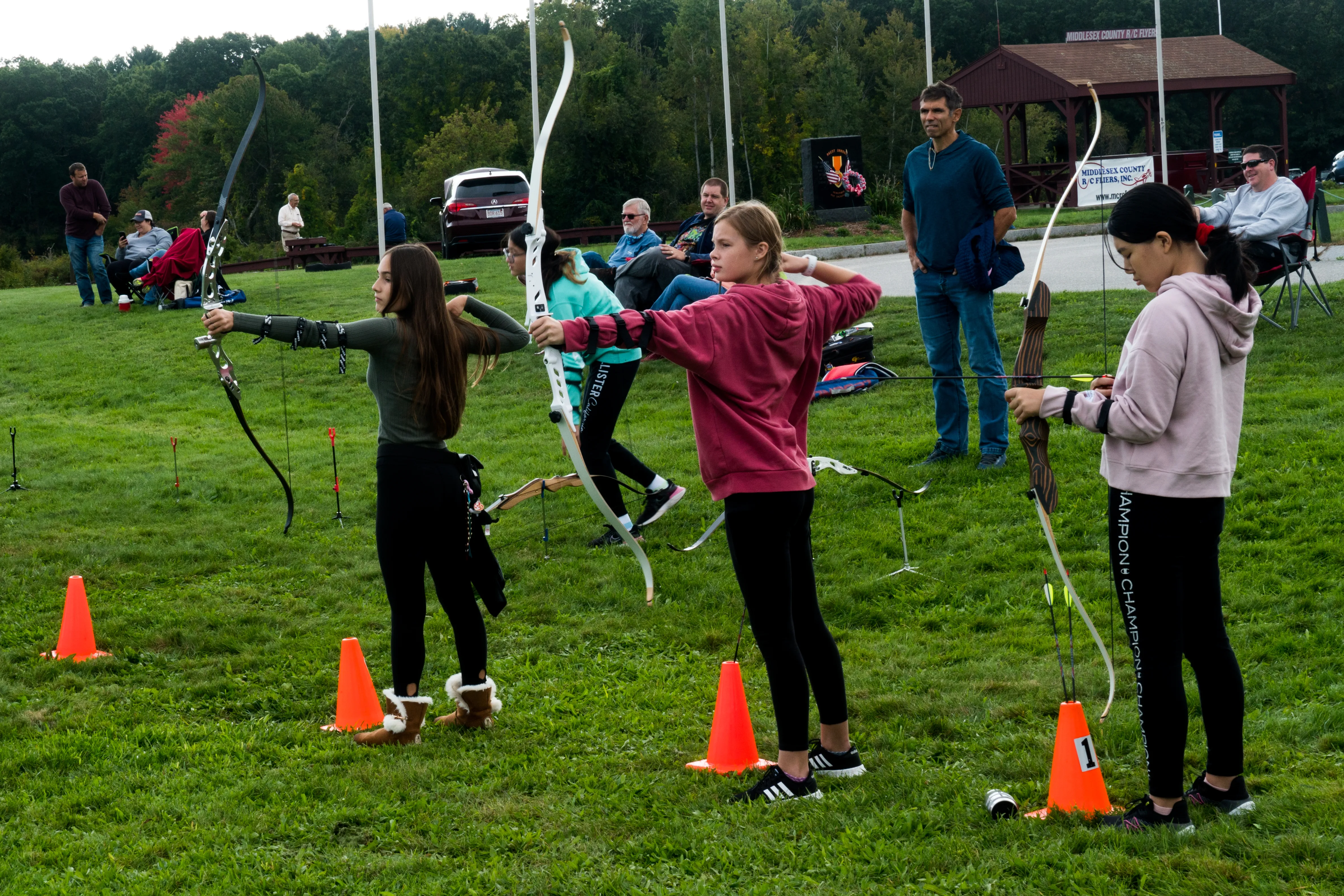 Group of teenage archers on a grass field, several drawing recurve bows simultaneously with trees in the background
