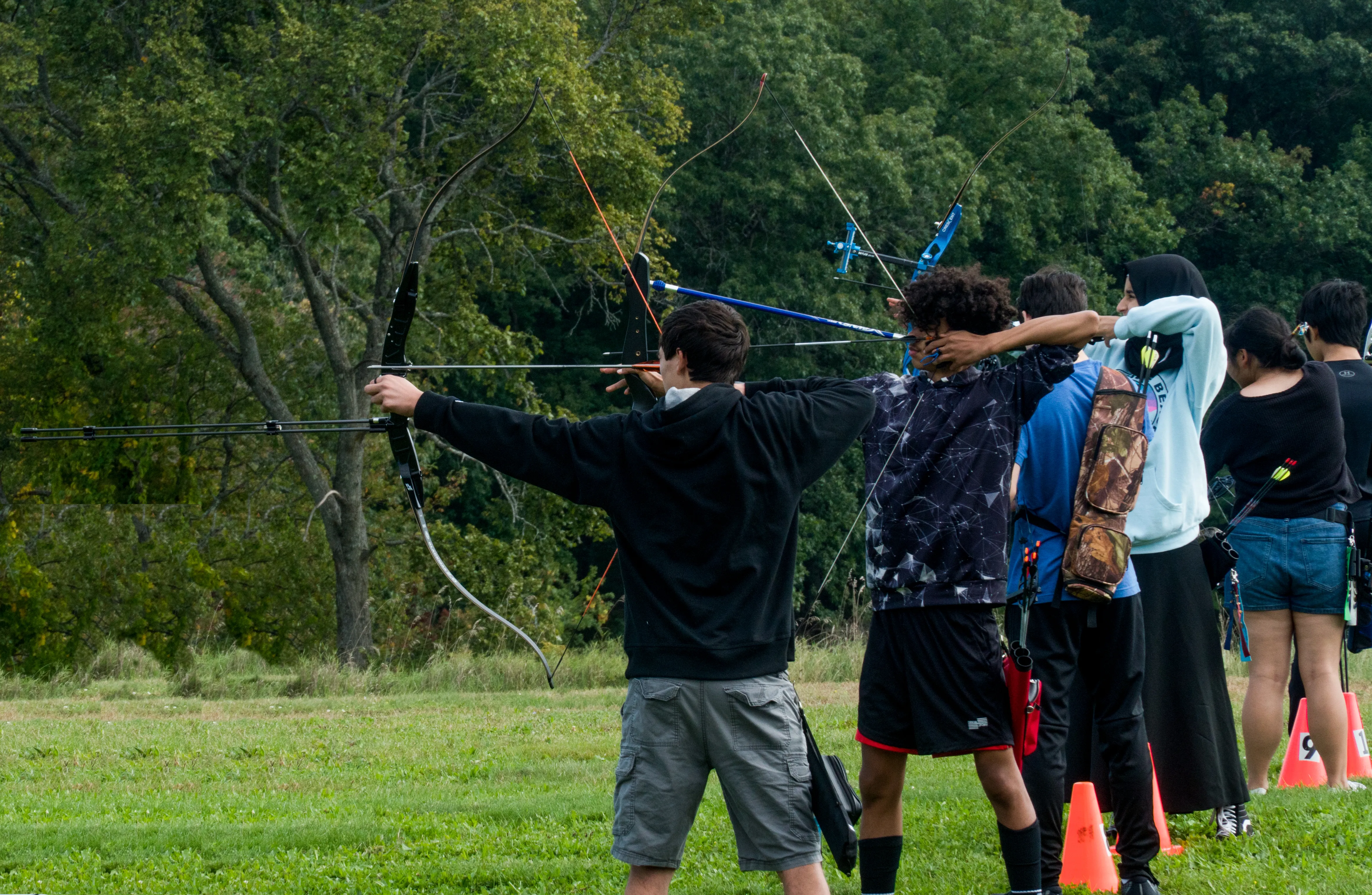 Adult instructor in blue shirt coaching young children with bows on a grass field, with archery targets lined up in the background